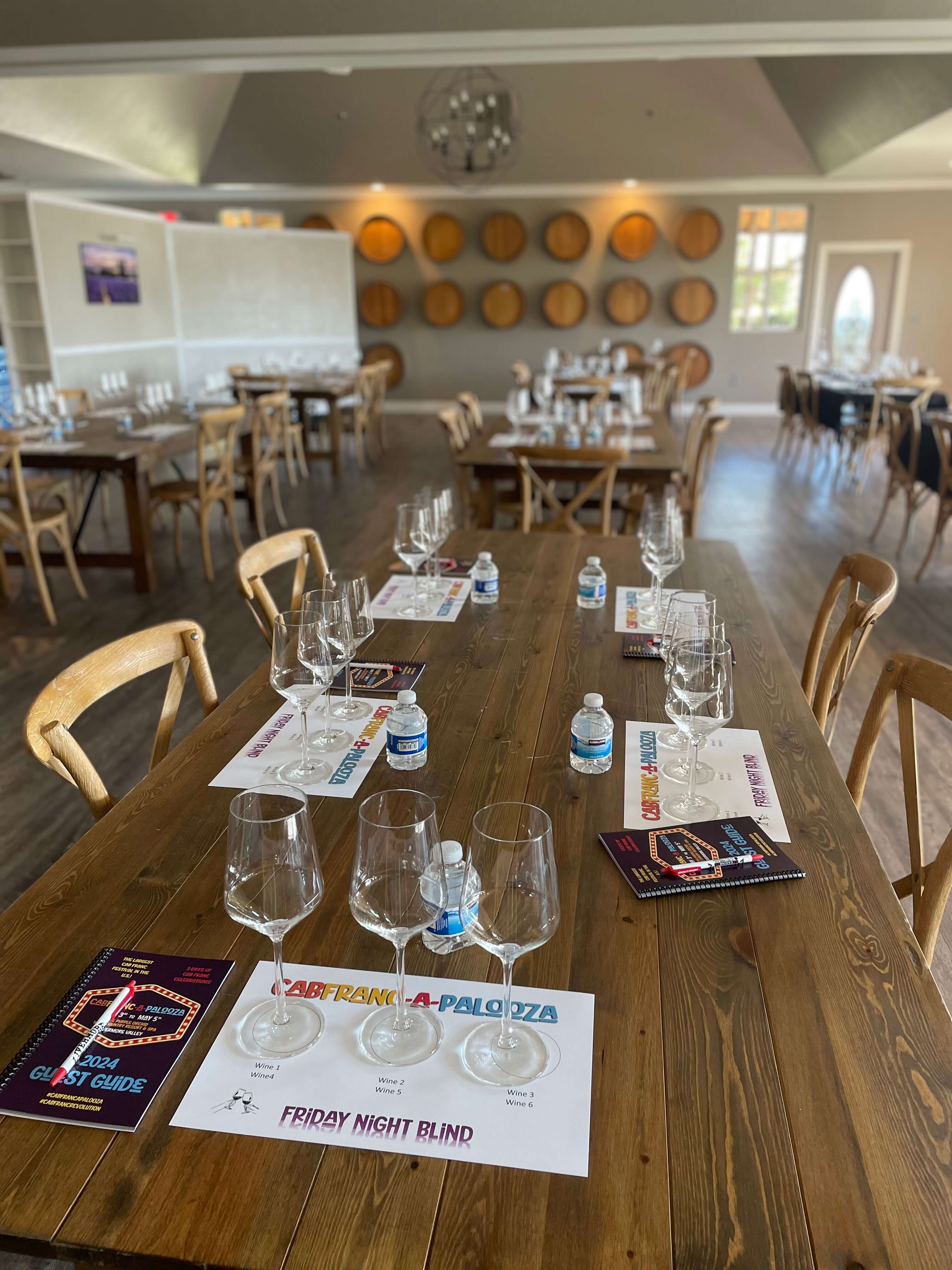 A beautifully arranged dining space set for a wine tasting event, featuring glasses and guest guides on each table.