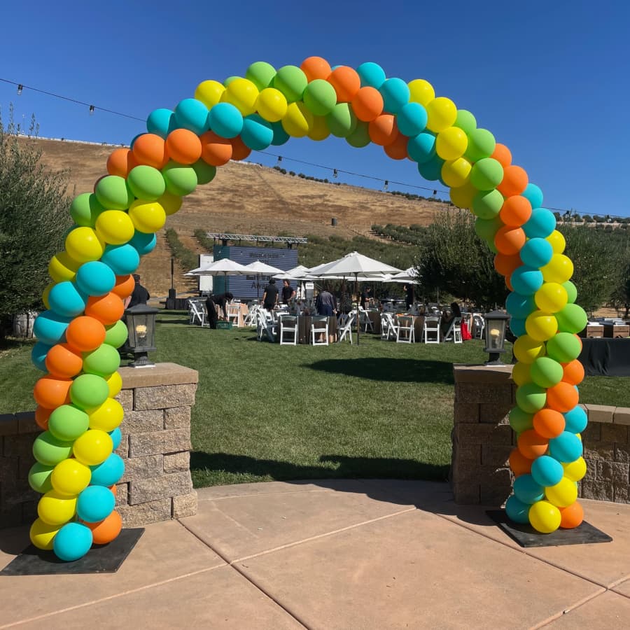 A vibrant balloon arch in orange, green, blue, and yellow stands in an outdoor event space with tables and tents.