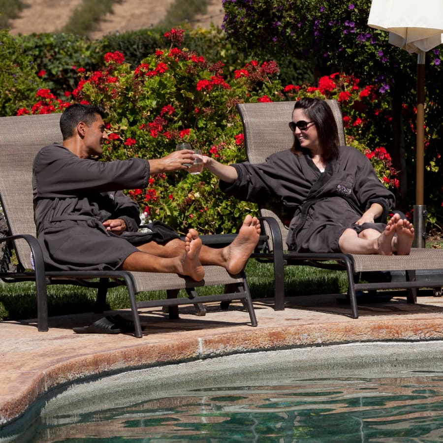 A man and woman in bathrobes clink glasses while lounging by a pool surrounded by colorful flowers.