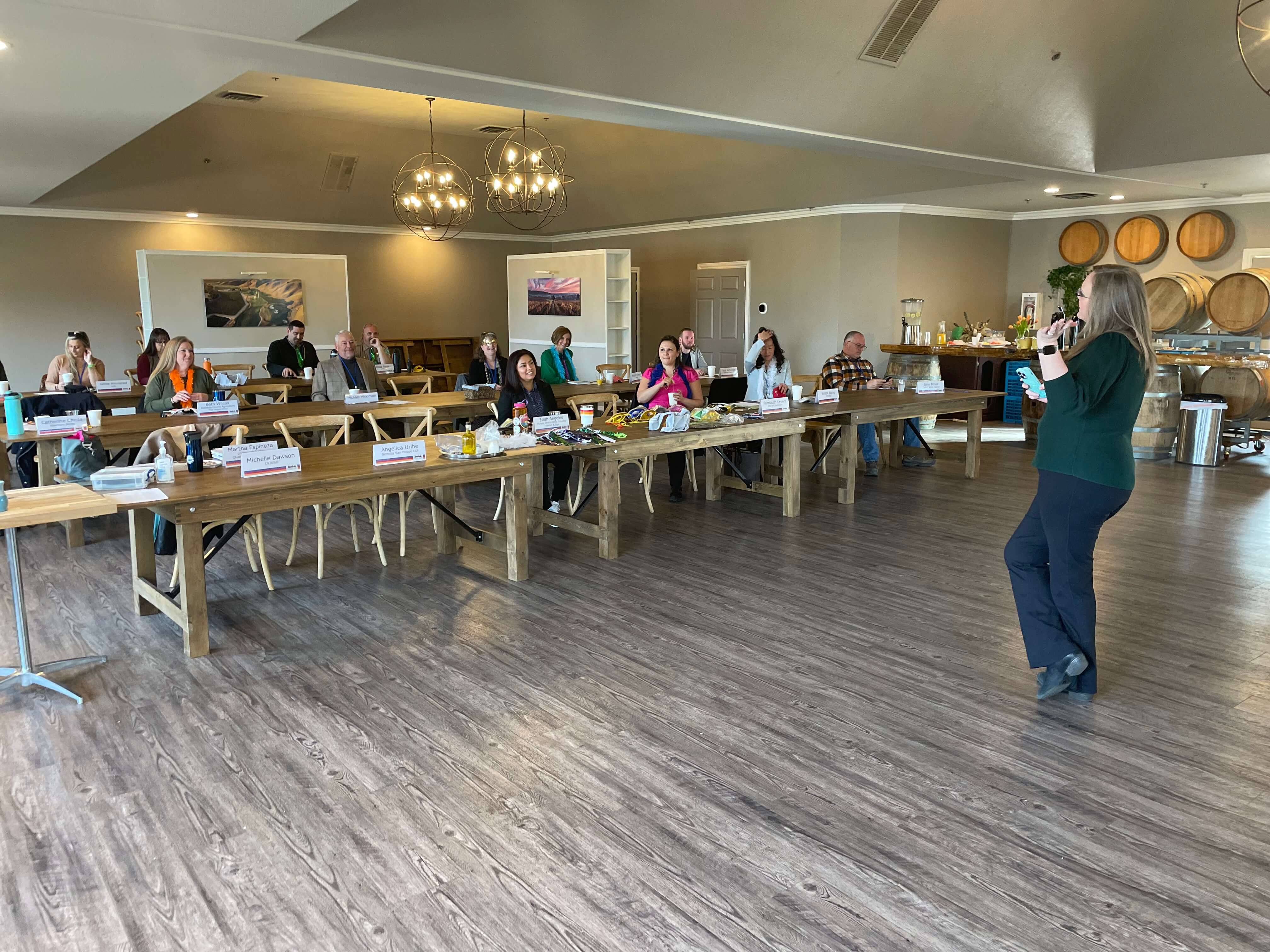A woman speaks at the front of a well-lit meeting room filled with seated audience members.
