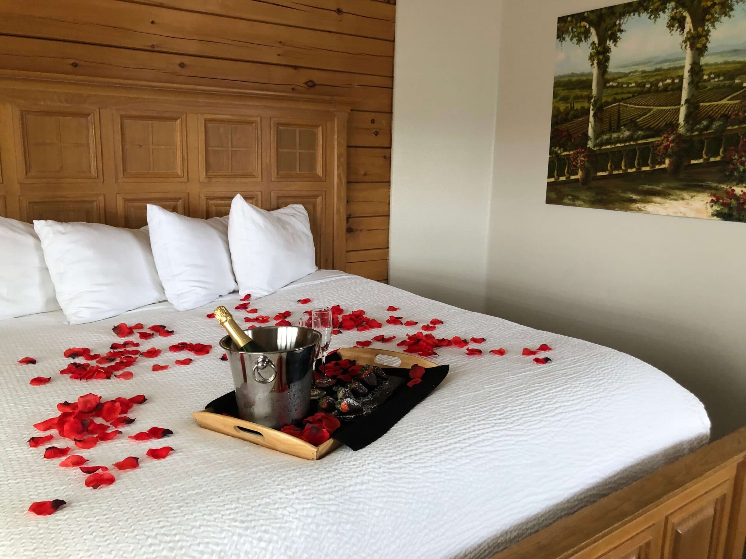 A romantic bedroom setup featuring a bed adorned with rose petals, champagne in an ice bucket, and a tray with chocolate-covered strawberries.