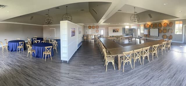 A spacious interior event space with round and rectangular tables, featuring wooden decor and large windows.