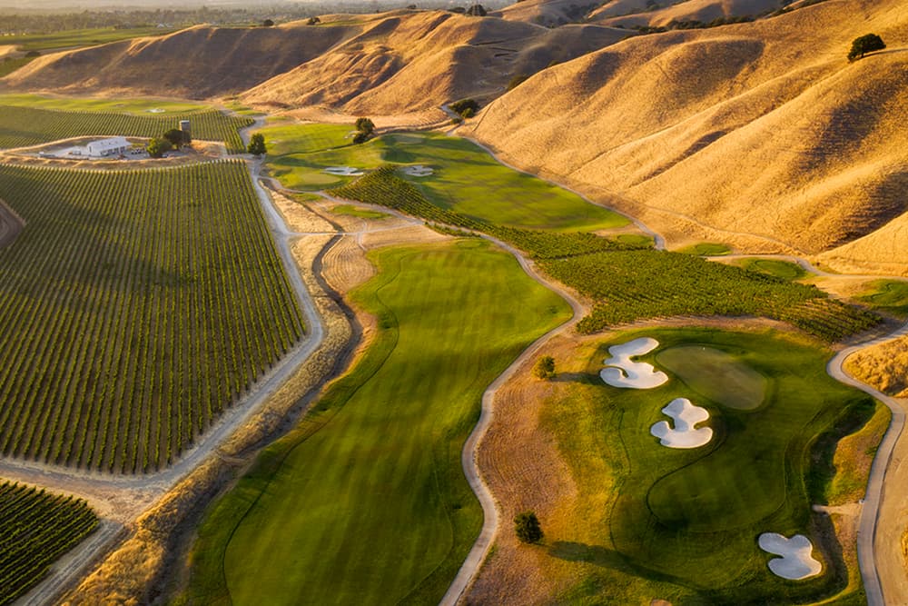 Aerial view of a golf course surrounded by rolling hills and vineyards.