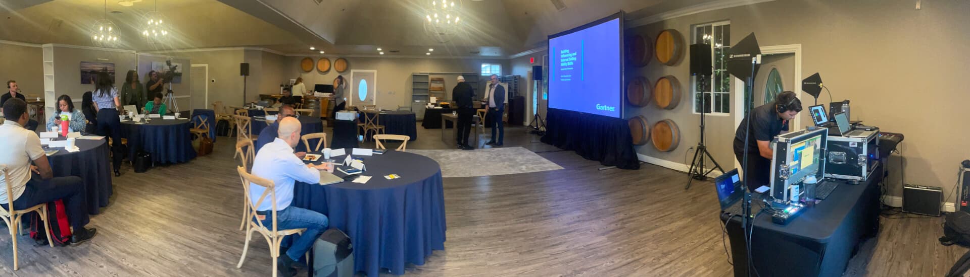 A spacious meeting room set up for a conference, featuring round tables, audiovisual equipment, and attendees engaged in discussions.