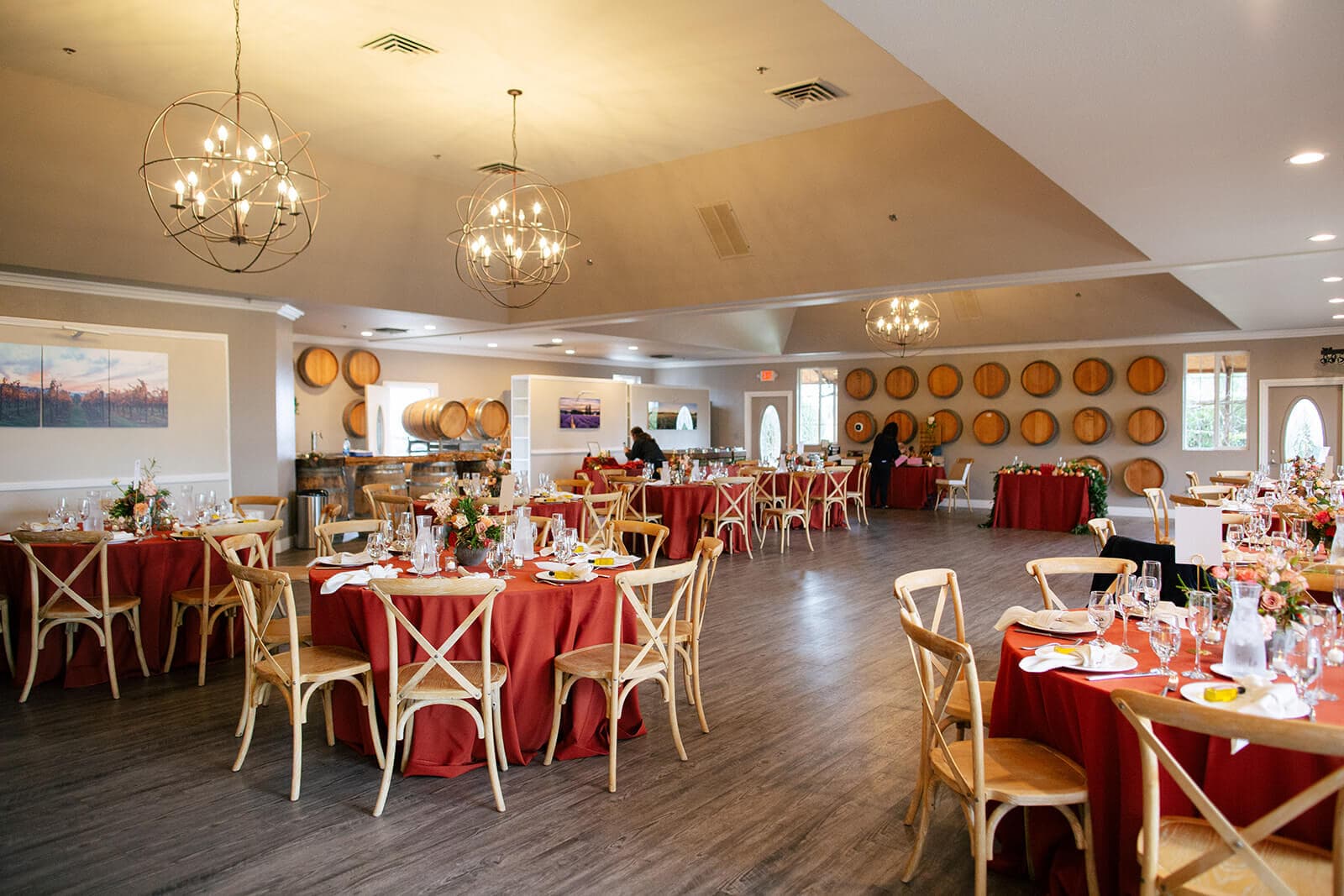 A spacious event hall set with red tablecloths, wooden chairs, and wine barrels, illuminated by stylish chandeliers.