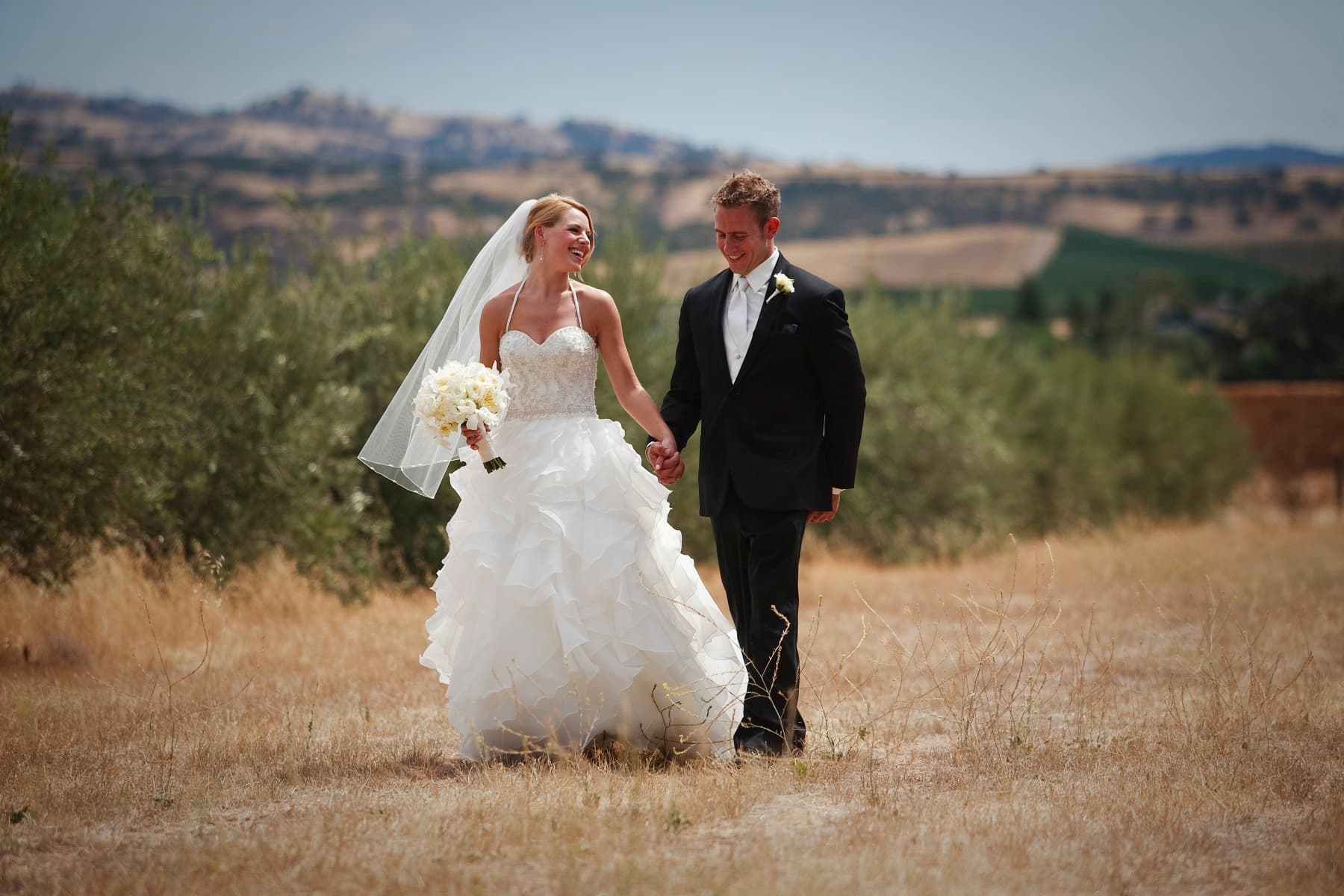 A bride and groom hold hands and walk together through a sunlit field.