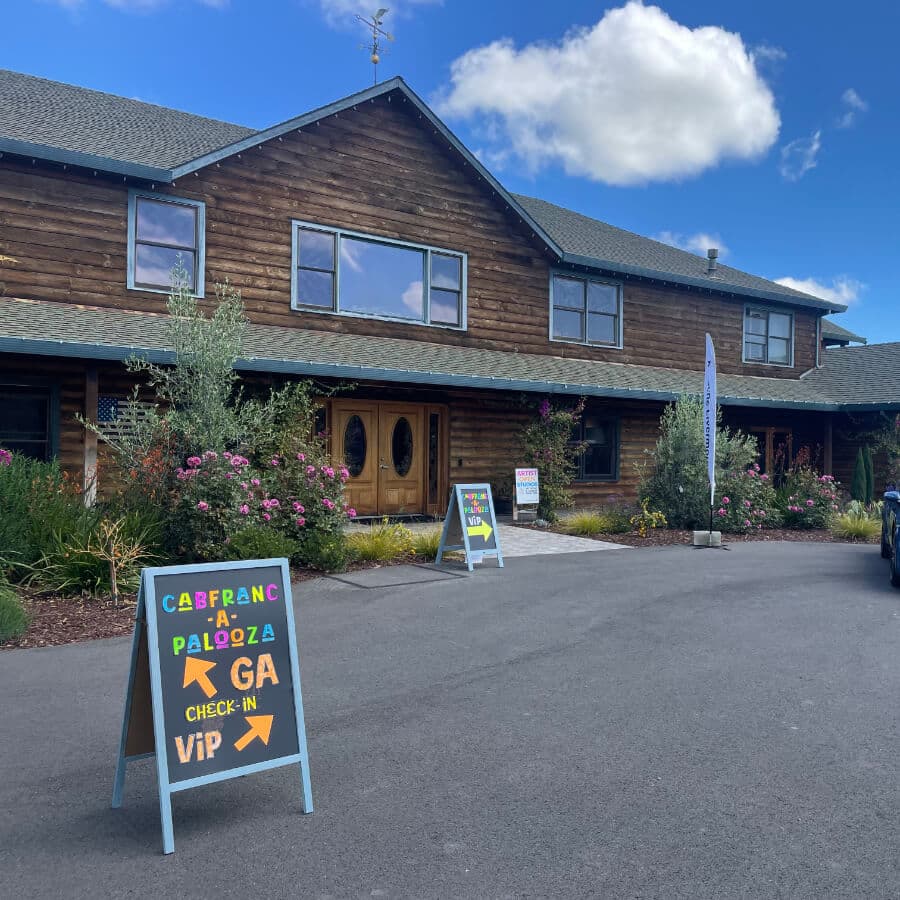 A rustic wooden building with event check-in signs and colorful flower landscaping under a partly cloudy sky.