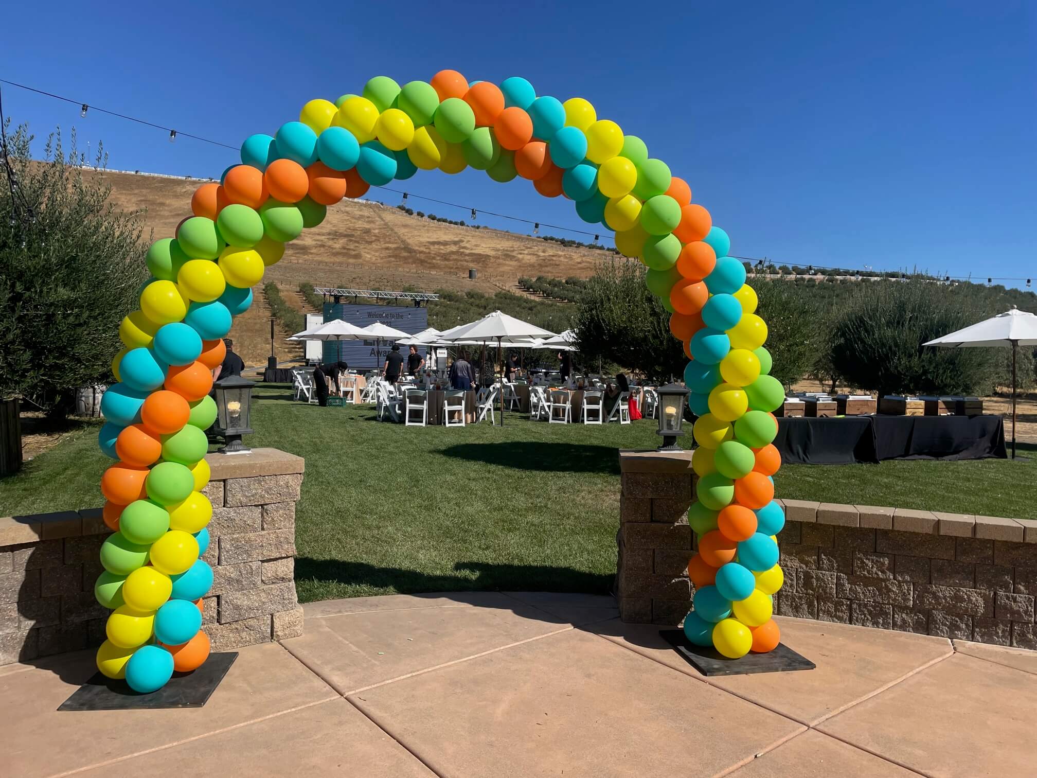 A vibrant balloon arch in blue, orange, yellow, and green stands at the entrance to an outdoor event space.