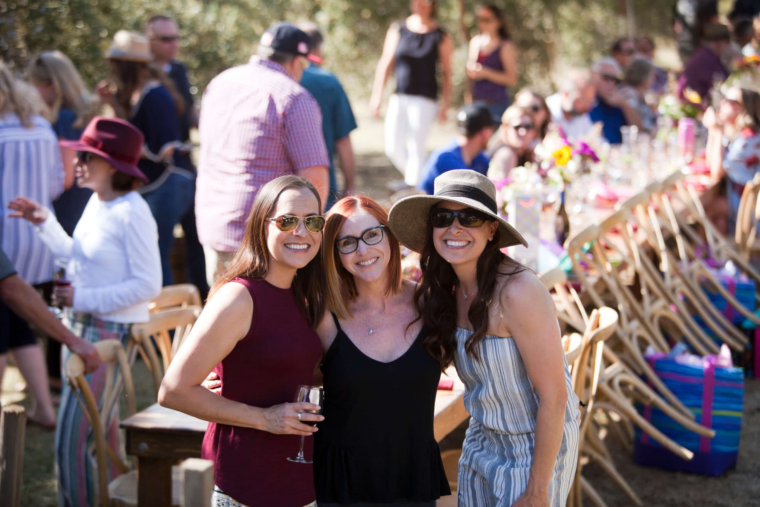 Three smiling women pose together at an outdoor gathering with a festive atmosphere.