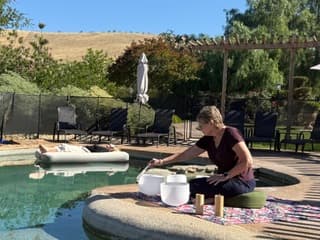 A woman sits by a pool, arranging crystal bowls and candles on a blanket while another person relaxes on a floating lounge nearby.