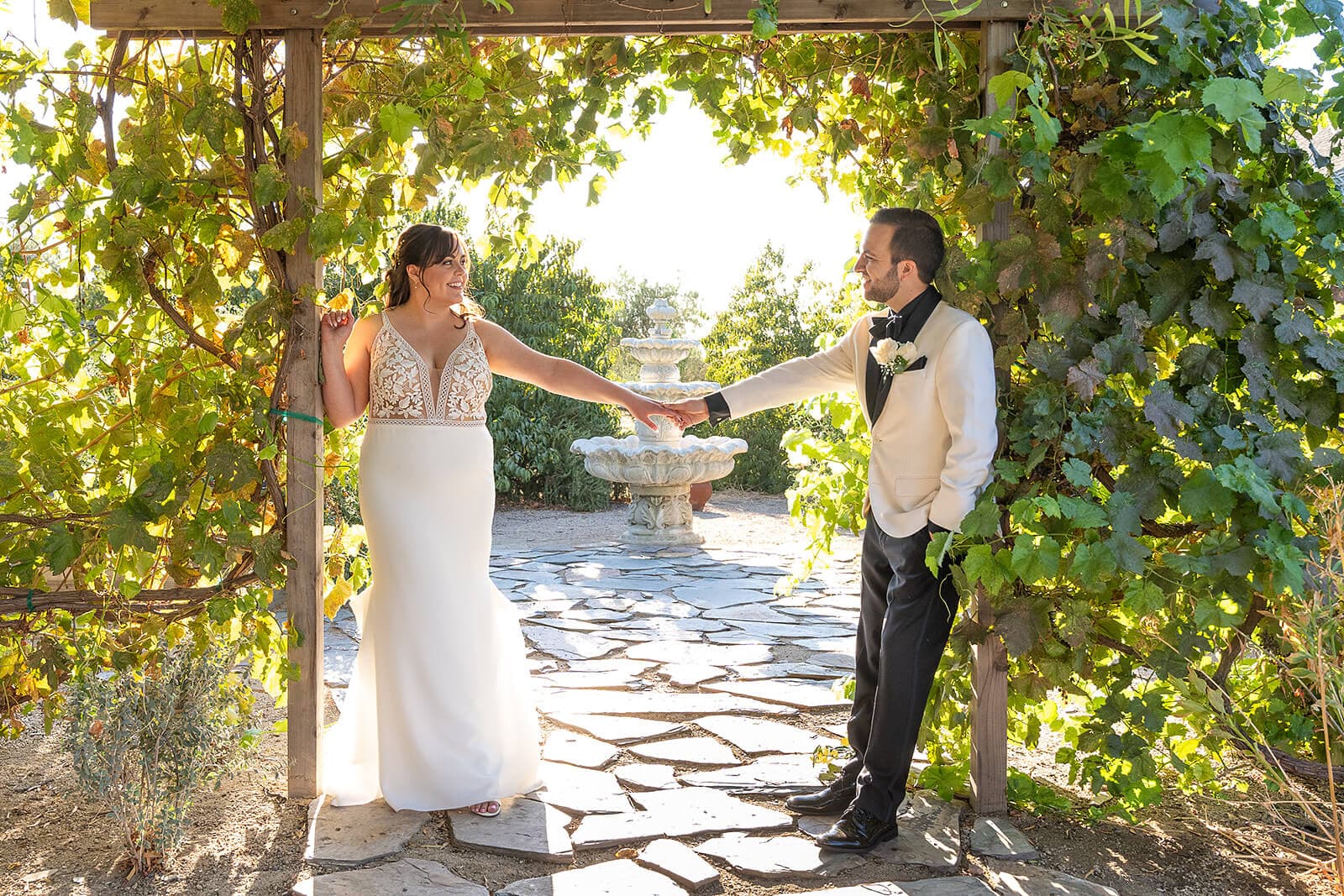 A bride and groom holding hands under a vine-covered arbor, surrounded by lush greenery and sunlight.