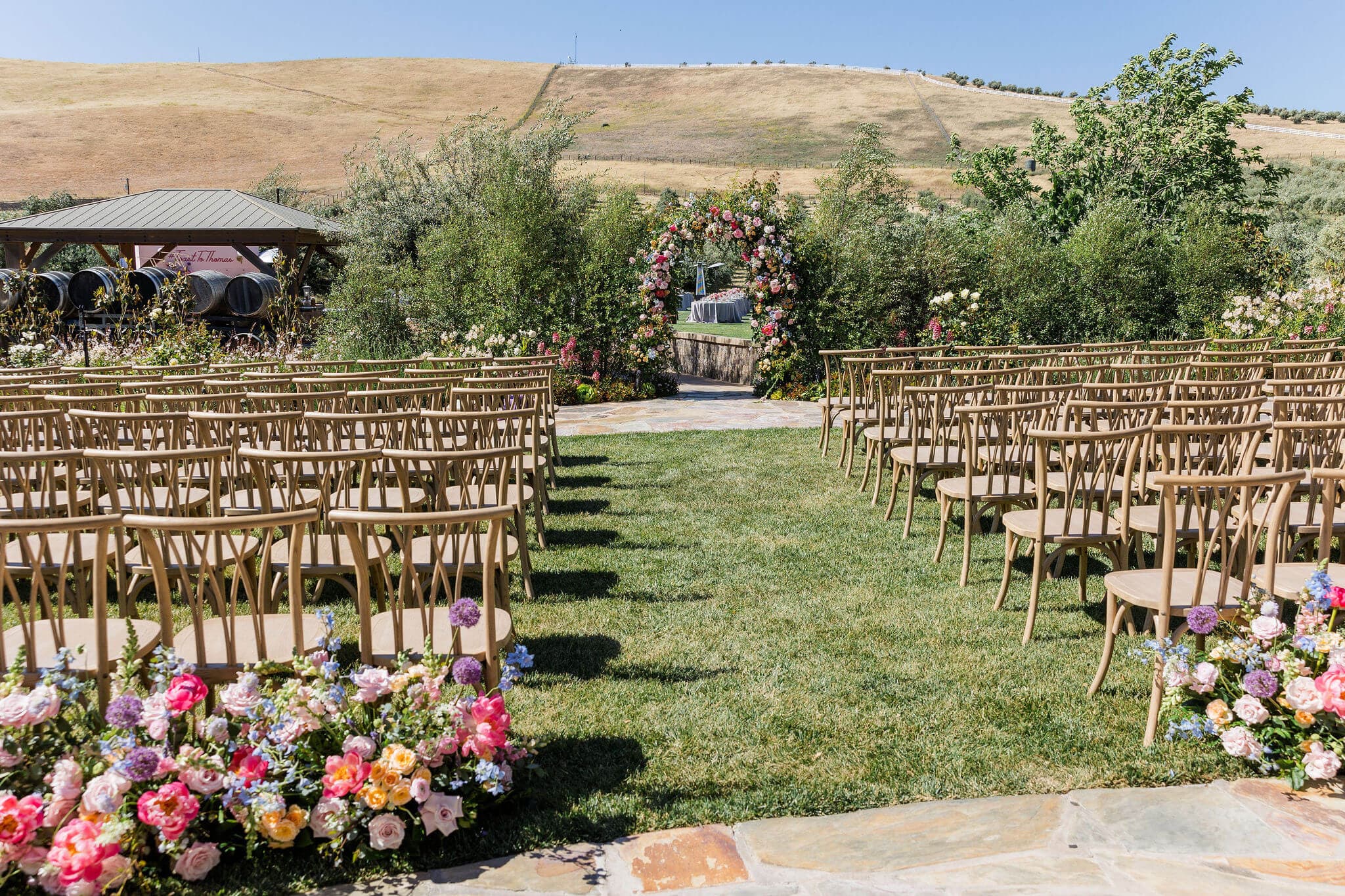 An outdoor wedding venue featuring rows of wooden chairs and a floral archway on a grassy area.