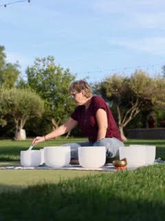 A woman sits on grass, playing crystal bowls during a sound bath session.