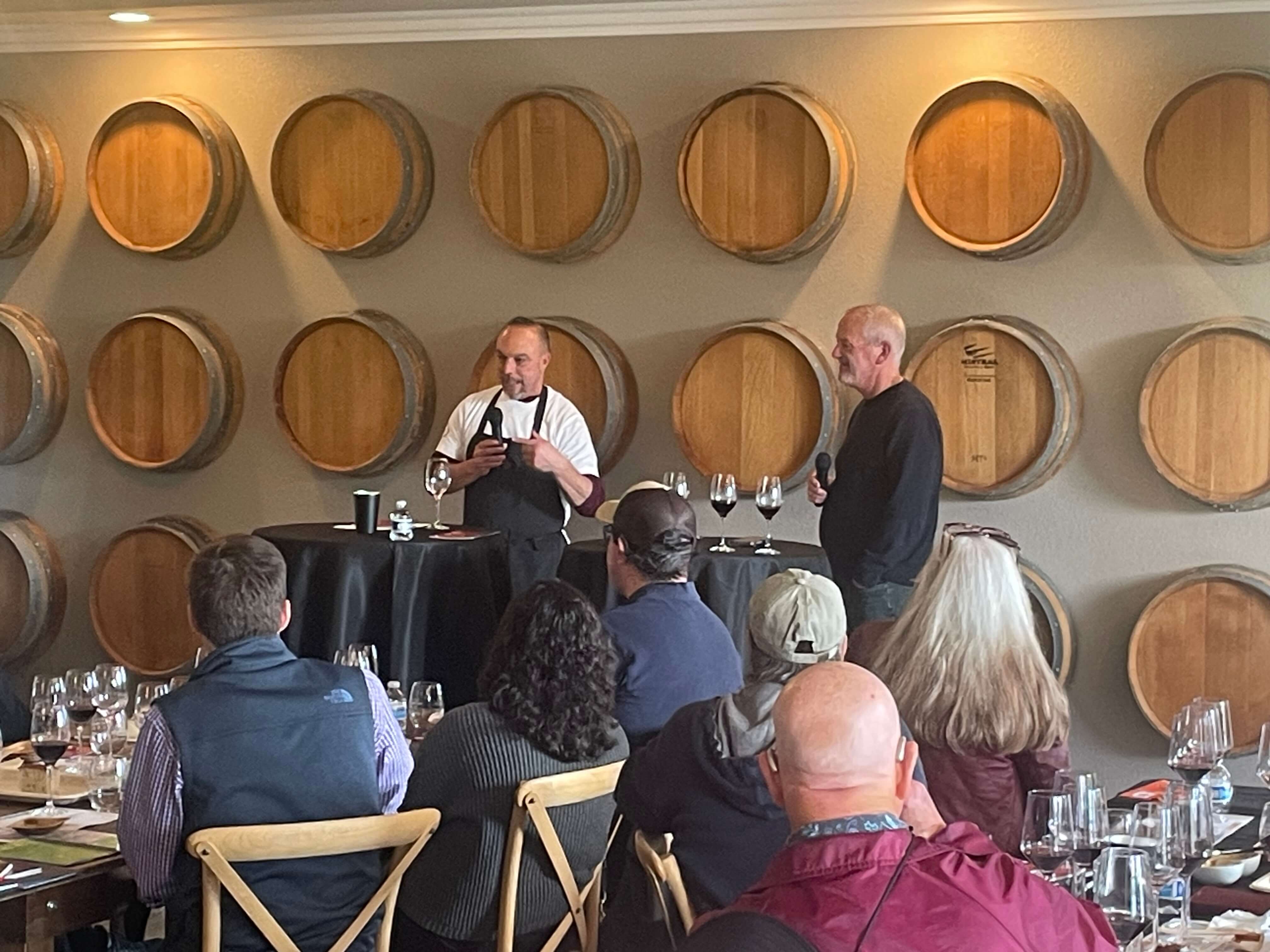 A speaker presents to an audience in front of wine barrels at a tasting event.