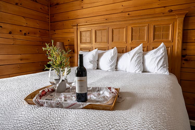 A cozy bedroom with a wooden headboard, featuring a neatly made bed, a tray with a bottle of wine, two glasses, and a small vase of greenery.