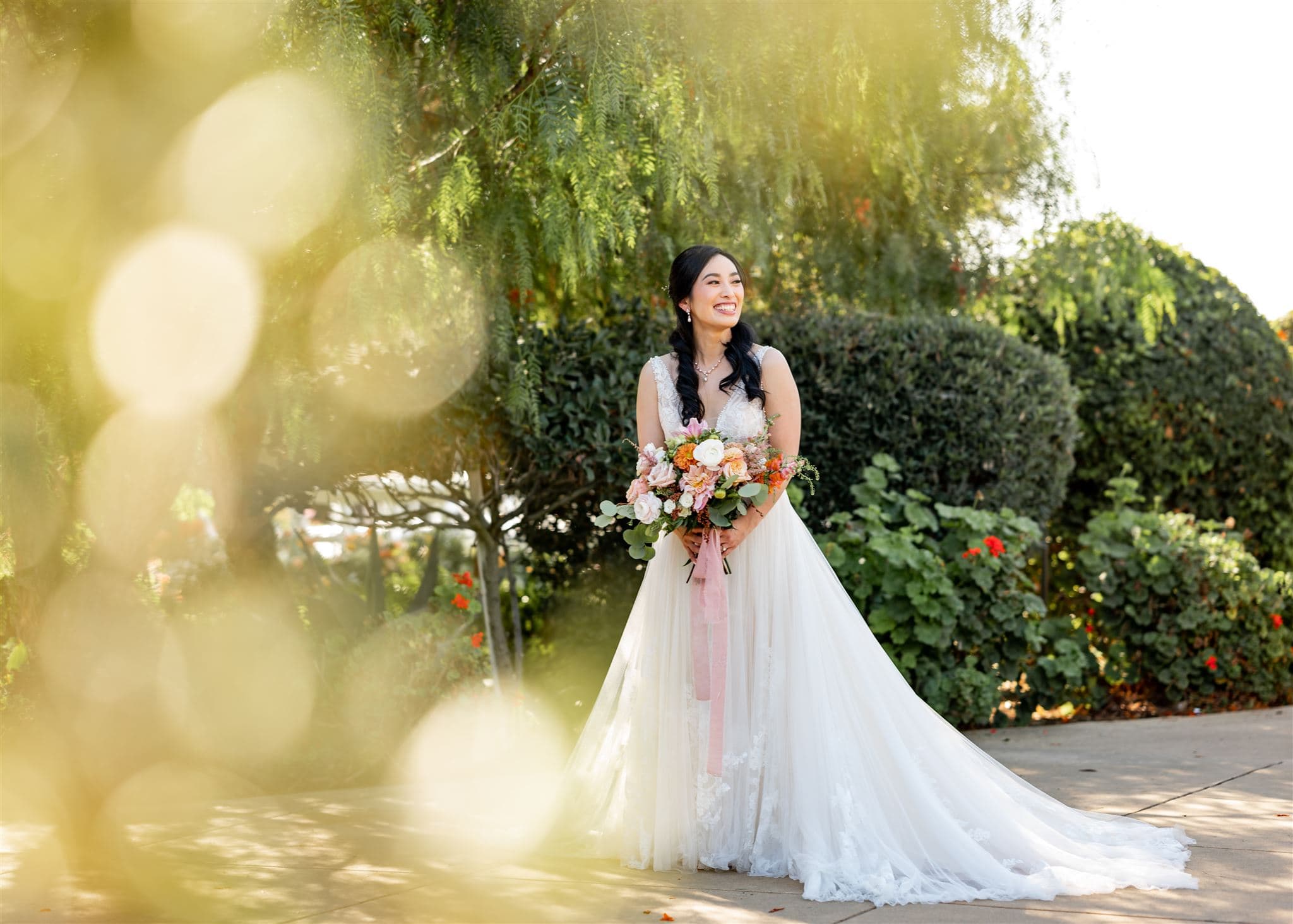 A smiling bride stands outdoors, holding a floral bouquet while wearing a flowing wedding gown.