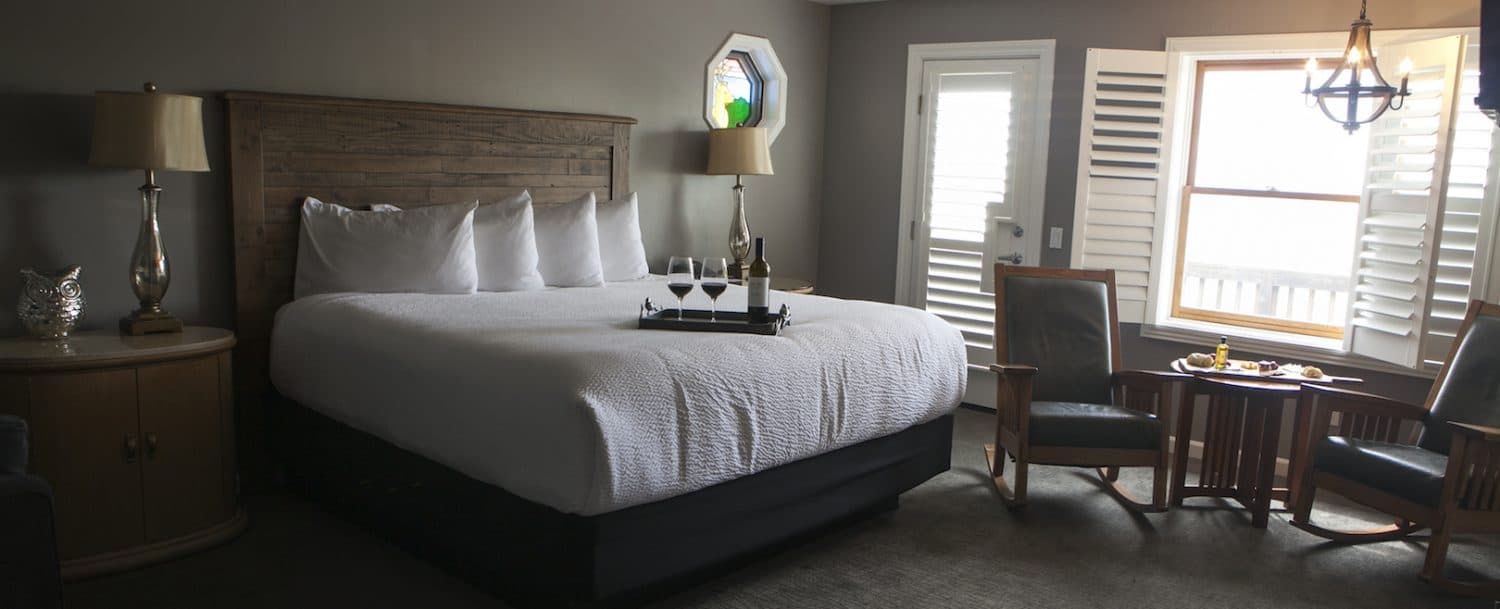 A serene room with a king-size bed, two armchairs, a window with shutters, and a tray with a bottle of wine on the bed.