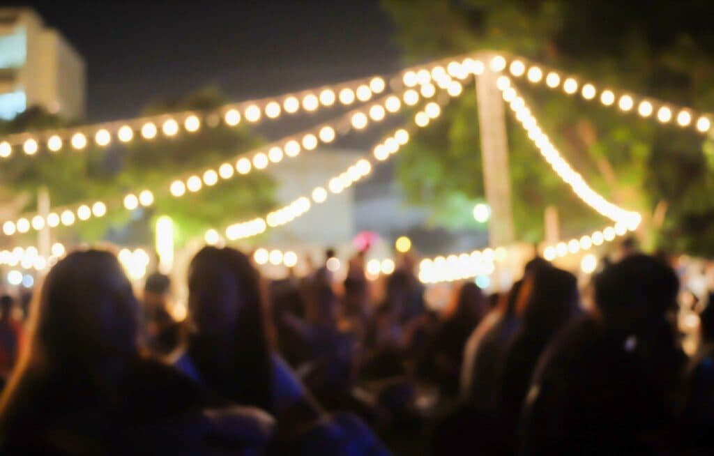 A group of people listing to an evening outdoor music event with string lights hanging above them A group of people listing to an evening outdoor music event with string lights hanging above them